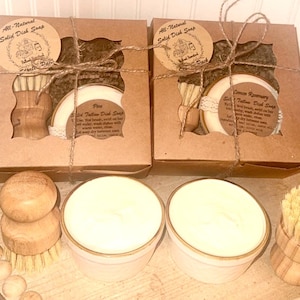 May include: Two boxed sets of solid dish soap with wooden dish brushes. Each box is tied with twine and features a round label with text. Two additional soaps sit in white ceramic containers. The text on the label reads "All-Natural Solid Dish Soap".