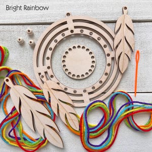 May include: A craft kit with the text "Bright Rainbow" includes wooden circular frames, feather shapes, beads, an orange needle, and colorful yarn in a rainbow pattern. The kit is designed for crafting and DIY projects.