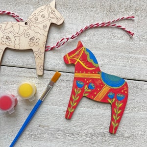 May include: Two wooden Dala horse ornaments, one painted red with blue and yellow details, the other unpainted with a floral design. Paint pots and a paintbrush are included.