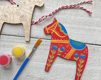 May include: Two wooden Dala horse ornaments, one painted red with blue and yellow details, the other unpainted with a floral design. Paint pots and a paintbrush are included.