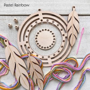 May include: A wooden craft kit with a pastel rainbow theme. The kit includes a circular frame, a smaller circular base, three feather-shaped cutouts, wooden beads, a pink needle, and colorful yarn. The text "Pastel Rainbow" is in the upper left corner.