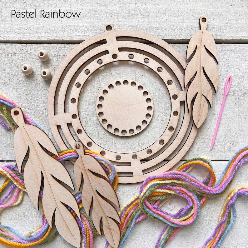 May include: A wooden craft kit with a pastel rainbow theme. The kit includes a circular frame, a smaller circular base, three feather-shaped cutouts, wooden beads, a pink needle, and colorful yarn. The text "Pastel Rainbow" is in the upper left corner.