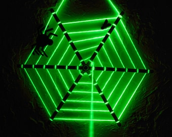 以下が含まれることがあります： 暗い背景に緑色のレーザー光が蜘蛛の巣のデザインを作り出しています。黒い蜘蛛と蝶のシルエットが巣の中にあります。