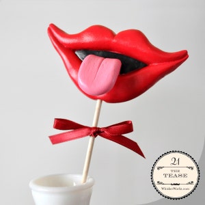 May include: A red and pink fondant mouth with a tongue sticking out, on a wooden stick with a red ribbon bow. The mouth is on a white pedestal. The text "21 The Tease WhisperWorks.com" is on a black and white circle.