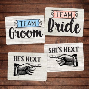 Op de afbeelding: Vier rechthoekige houten borden met tekst voor een bruiloft. De borden luiden "Team Groom", "Team Bride", "He's Next" en "She's Next". De borden hebben een houtnerf achtergrond en zwarte tekst.
