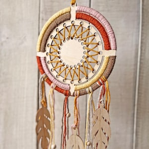 May include: A handcrafted dreamcatcher with a wooden frame and intricate woven design. The circular frame is wrapped in layers of yarn in shades of brown, orange, pink, and yellow. Wooden leaf-shaped pendants and beads hang from the bottom, adding a natural touch.