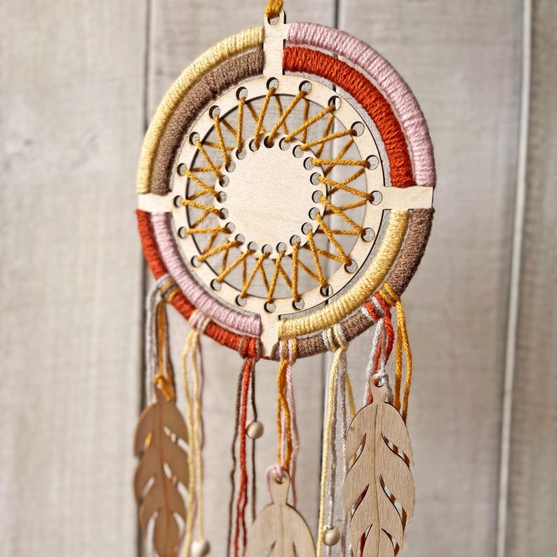 May include: A handcrafted dreamcatcher with a wooden frame and intricate woven design. The circular frame is wrapped in layers of yarn in shades of brown, orange, pink, and yellow. Wooden leaf-shaped pendants and beads hang from the bottom, adding a natural touch.