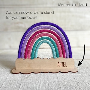 Pu&ograve; includere: Un arcobaleno decorativo con una base in legno. L'arcobaleno presenta sfumature di viola, rosa e turchese. La base in legno ha un bordo smerlato e il nome "ARIEL" &egrave; inciso. Il testo sull'immagine dice: "Puoi ordinare un supporto per il tuo arcobaleno!"