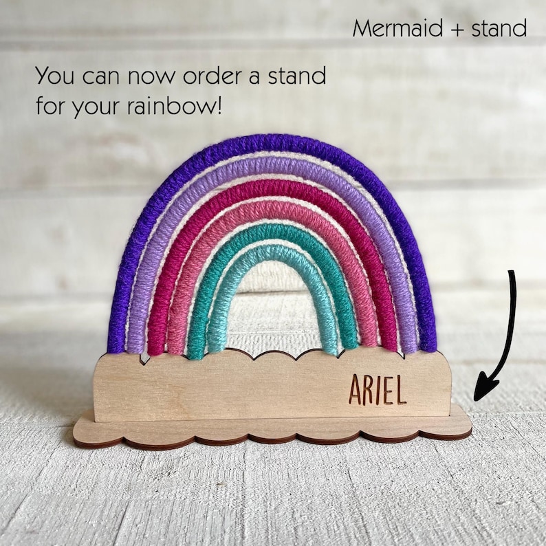 Pu&ograve; includere: Un arcobaleno decorativo con una base in legno. L'arcobaleno presenta sfumature di viola, rosa e turchese. La base in legno ha un bordo smerlato e il nome "ARIEL" &egrave; inciso. Il testo sull'immagine dice: "Puoi ordinare un supporto per il tuo arcobaleno!"