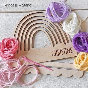 Pu&ograve; includere: Artigianato in legno a forma di arcobaleno con il nome "CHRISTINE" inciso sulla base a nuvola. L'arcobaleno &egrave; circondato da gomitoli di lana rosa, viola, bianca e gialla, insieme a un ago per maglieria rosa e un supporto in legno.