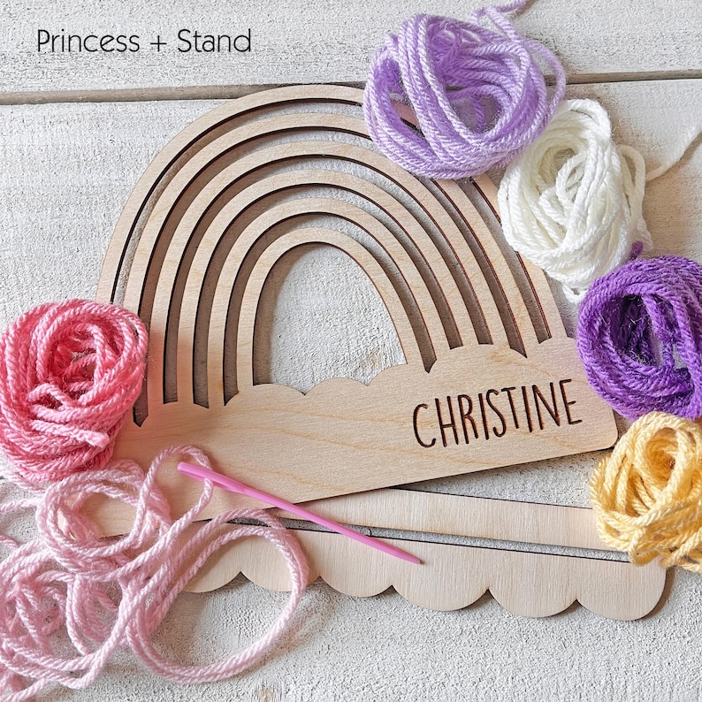 Pu&ograve; includere: Artigianato in legno a forma di arcobaleno con il nome "CHRISTINE" inciso sulla base a nuvola. L'arcobaleno &egrave; circondato da gomitoli di lana rosa, viola, bianca e gialla, insieme a un ago per maglieria rosa e un supporto in legno.