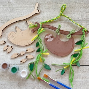 May include: A wooden sloth hanging mobile craft kit with green and yellow yarn, brown paint, and two paintbrushes. The kit includes a sloth hanging from a branch, leaves, and a sloth on a separate piece of wood.