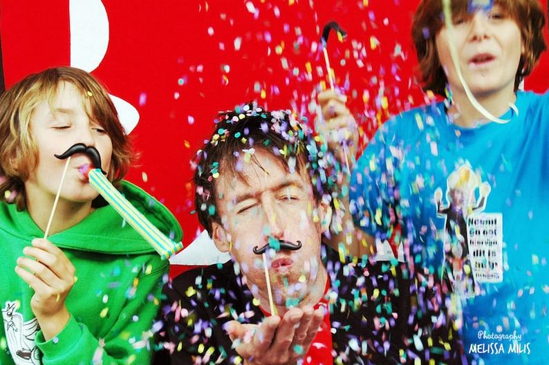 May include: Three people are celebrating with confetti. The person in the middle is blowing confetti out of their hand. The person on the left is wearing a green hoodie and blowing into a party favor. The person on the right is wearing a blue shirt and holding a party favor.