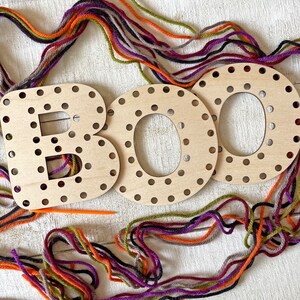 以下が含まれることがあります： 糸を通す穴のある「BOO」の文字が書かれた木製の文字で、オレンジ、紫、緑、グレーのカラフルな糸で囲まれています。オレンジ色の糸通し針が見えます。文字は明るい色の木でできており、ハロウィーンのクラフトプロジェクトに最適です。