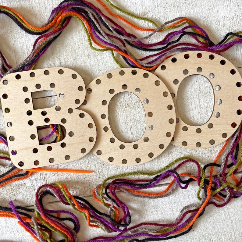 以下が含まれることがあります： 糸を通す穴のある「BOO」の文字が書かれた木製の文字で、オレンジ、紫、緑、グレーのカラフルな糸で囲まれています。オレンジ色の糸通し針が見えます。文字は明るい色の木でできており、ハロウィーンのクラフトプロジェクトに最適です。