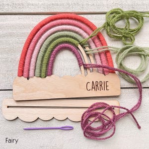Pu&ograve; includere: Decorazione murale a forma di arcobaleno fatta a mano con base in legno. L'arcobaleno presenta fili rossi, rosa e verdi. La base in legno ha inciso il nome "CARRIE". Include filo e un ago viola. La parola "Fairy" &egrave; in basso.