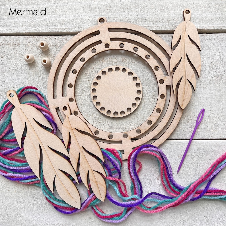 May include: A wooden dream catcher kit with a circular frame, feather-shaped cutouts, and colorful yarn in shades of teal, purple, and pink. Includes a needle and small wooden beads. The word "Mermaid" is in the top left corner.