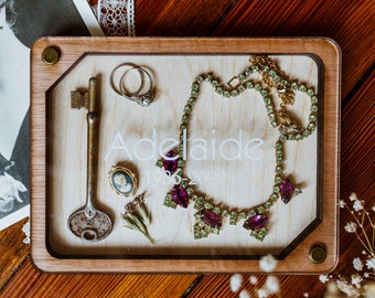Könnte beinhalten: Eine Holzkiste mit einem Glaseinsatz, der einen alten Schlüssel, zwei Ringe, ein kleines Kamee, ein paar getrocknete Blumen und eine grüne und violette Strasskette enthält. Die Kiste ist mit dem Namen "Adelaide" und den Daten "1936-2022" graviert.
