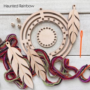 May include: A wooden dream catcher kit with the text "Haunted Rainbow". The kit includes a circular frame, a smaller circular piece, wooden feather cutouts, wooden beads, colorful yarn, and an orange needle. The components are arranged on a white wooden surface.