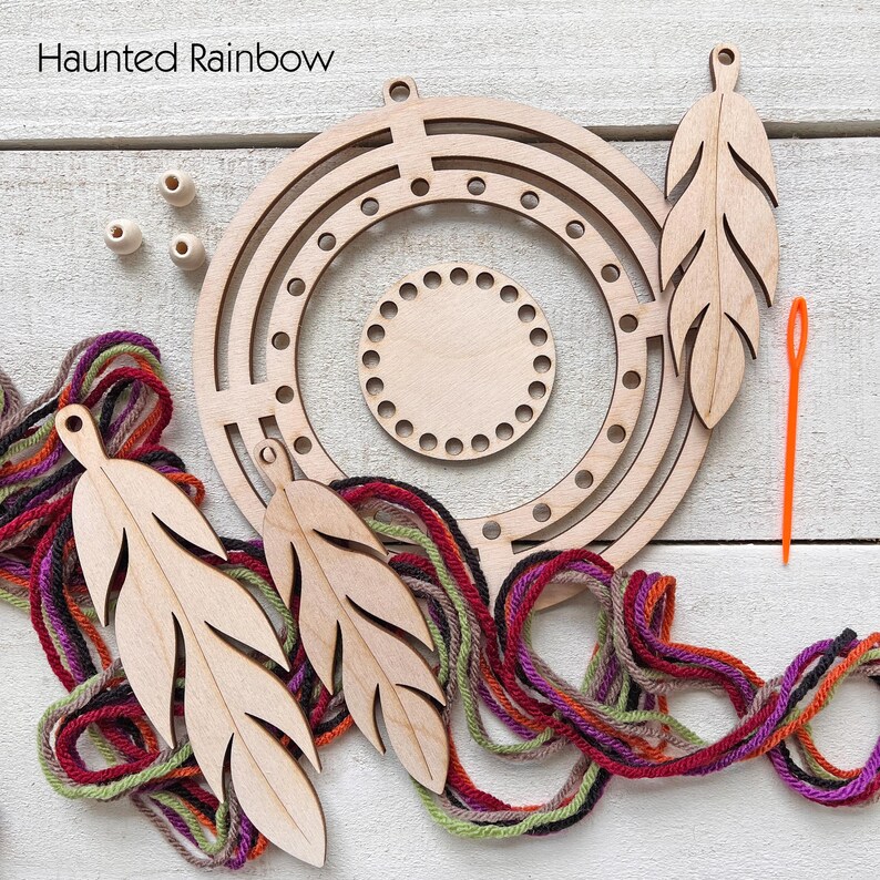 May include: A wooden dream catcher kit with the text "Haunted Rainbow". The kit includes a circular frame, a smaller circular piece, wooden feather cutouts, wooden beads, colorful yarn, and an orange needle. The components are arranged on a white wooden surface.