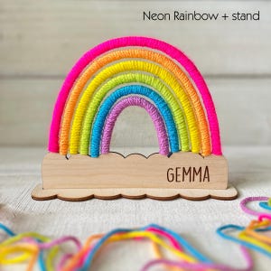 Pu&ograve; includere: Un ornamento decorativo a forma di arcobaleno con una base in legno. L'arcobaleno presenta colori neon vibranti, tra cui rosa, arancione, giallo, verde, blu e viola. La base in legno &egrave; incisa con il nome "GEMMA" e ha un bordo smerlato. Il testo "Neon Rainbow + stand" &egrave; in alto.