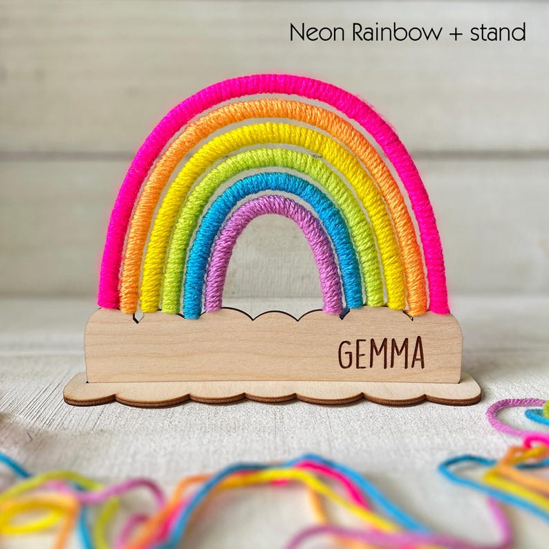 Pu&ograve; includere: Un ornamento decorativo a forma di arcobaleno con una base in legno. L'arcobaleno presenta colori neon vibranti, tra cui rosa, arancione, giallo, verde, blu e viola. La base in legno &egrave; incisa con il nome "GEMMA" e ha un bordo smerlato. Il testo "Neon Rainbow + stand" &egrave; in alto.
