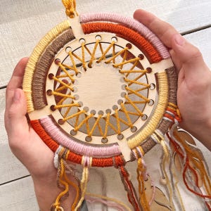 May include: A handmade dreamcatcher with a wooden frame and colorful yarn in shades of yellow, brown, orange, and pink. The center features a wooden circle with a woven pattern. Decorative feathers hang from the bottom.