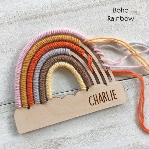 Pu&ograve; includere: Decorazione in legno a forma di arcobaleno con base a forma di nuvola. L'arcobaleno presenta strati di filato rosa, giallo, arancione, grigio e marrone. Il nome "CHARLIE" &egrave; inciso sulla base. Il testo "Boho Rainbow" &egrave; in alto a destra.