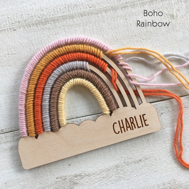 Pu&ograve; includere: Decorazione in legno a forma di arcobaleno con base a forma di nuvola. L'arcobaleno presenta strati di filato rosa, giallo, arancione, grigio e marrone. Il nome "CHARLIE" &egrave; inciso sulla base. Il testo "Boho Rainbow" &egrave; in alto a destra.