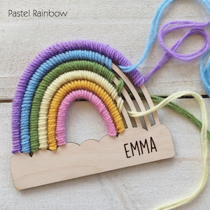Pu&ograve; includere: Un'applique a forma di arcobaleno pastello con una base a nuvola in legno. L'arcobaleno presenta strati di filato viola, blu, verde, giallo e rosa. Il nome "EMMA" &egrave; inciso sulla base della nuvola. Il testo "Pastel Rainbow" &egrave; in alto a sinistra.