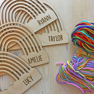 Pu&ograve; includere: Etichette in legno a forma di arcobaleno con i nomi "Ryann", "Taylor", "Amelie" e "Lucy" incisi. Una palla di filo colorato &egrave; accanto alle etichette. Le etichette sono in legno chiaro.