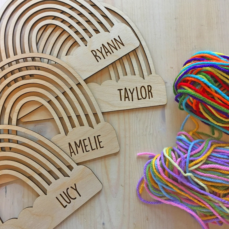 Pu&ograve; includere: Etichette in legno a forma di arcobaleno con i nomi "Ryann", "Taylor", "Amelie" e "Lucy" incisi. Una palla di filo colorato &egrave; accanto alle etichette. Le etichette sono in legno chiaro.