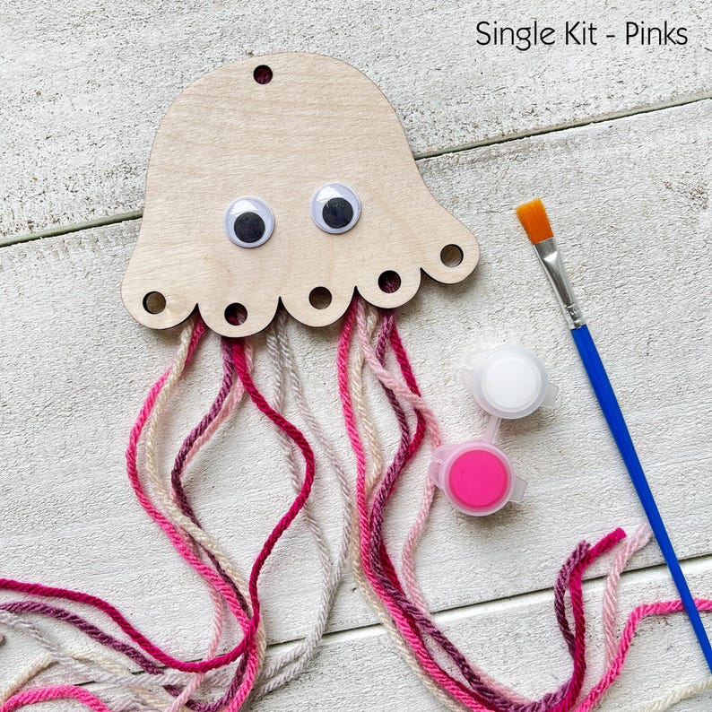 Puede incluir: Un kit de manualidades de medusas de madera con ojos saltones y tent&aacute;culos de hilo rosa, blanco y gris. Incluye botes de pintura y un pincel. El texto "Single Kit - Pinks" es visible.