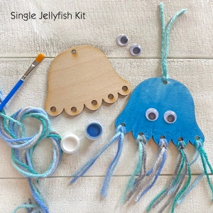 Puede incluir: Un kit de manualidades para una medusa individual. El kit incluye una forma de medusa de madera, pintura azul y blanca, un pincel, ojos saltones y lana en tonos azules, grises y blancos. El texto "Single Jellyfish Kit" es visible.
