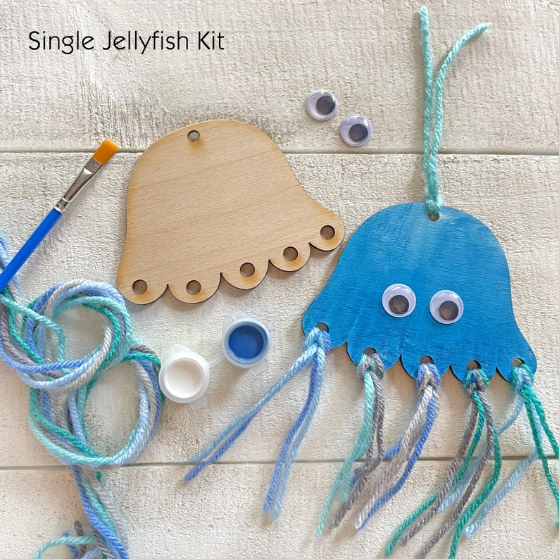 Puede incluir: Un kit de manualidades para una medusa individual. El kit incluye una forma de medusa de madera, pintura azul y blanca, un pincel, ojos saltones y lana en tonos azules, grises y blancos. El texto "Single Jellyfish Kit" es visible.