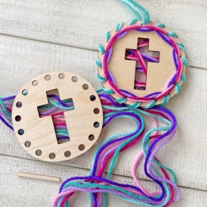 May include: Two wooden cross ornaments with colorful yarn. One is partially stitched with pink, purple, and teal yarn. The other is a blank wooden circle with a cross cutout and holes for stitching. A small wooden stick is also present.