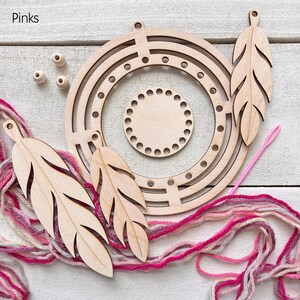 May include: A wooden craft kit with a circular frame, feather-shaped cutouts, and a small circular base. Pink and white yarn, wooden beads, and a pink needle are also included. The word "Pinks" is in the top left corner.