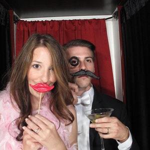Op de afbeelding: Een stel in een fotohokje met feestattributen. De vrouw houdt een rode plastic lippenprop vast en de man draagt een zwarte monocle en een nep snor. Ze lachen allebei.