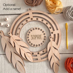 May include: A wooden rainbow-shaped craft kit with the name "Sophie" engraved on a circular piece. The kit includes wooden feathers, beads, and yarn in various colors. The text "Optional: Add a name!" is also visible.