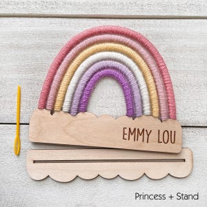 Pu&ograve; includere: Un'applique a forma di arcobaleno fatta a mano con base e supporto in legno. L'arcobaleno presenta strati di filato rosa, giallo, bianco e viola. La base in legno &egrave; incisa con il nome "EMMY LOU". Un ago giallo a sinistra.