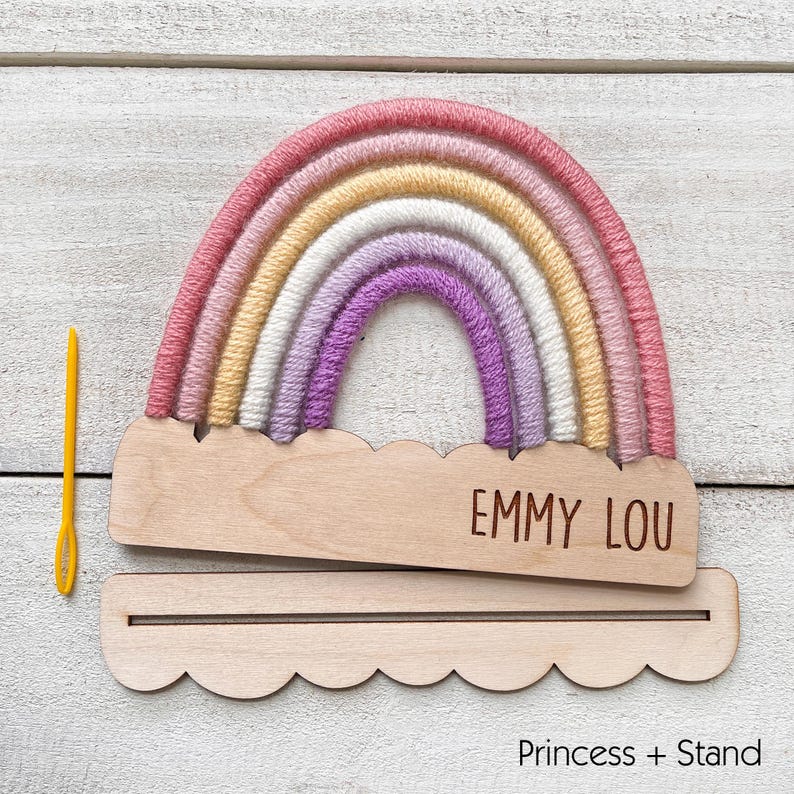 Pu&ograve; includere: Un'applique a forma di arcobaleno fatta a mano con base e supporto in legno. L'arcobaleno presenta strati di filato rosa, giallo, bianco e viola. La base in legno &egrave; incisa con il nome "EMMY LOU". Un ago giallo a sinistra.