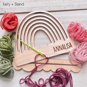 Pu&ograve; includere: Artigianato in legno a forma di arcobaleno con il nome "ANNALISA" inciso sulla base. Gomitoli di lana rosa, verde e rossa circondano l'oggetto, con un ago verde infilato con filo rosa. Il testo "Fairy + Stand" &egrave; nell'angolo in alto a sinistra.