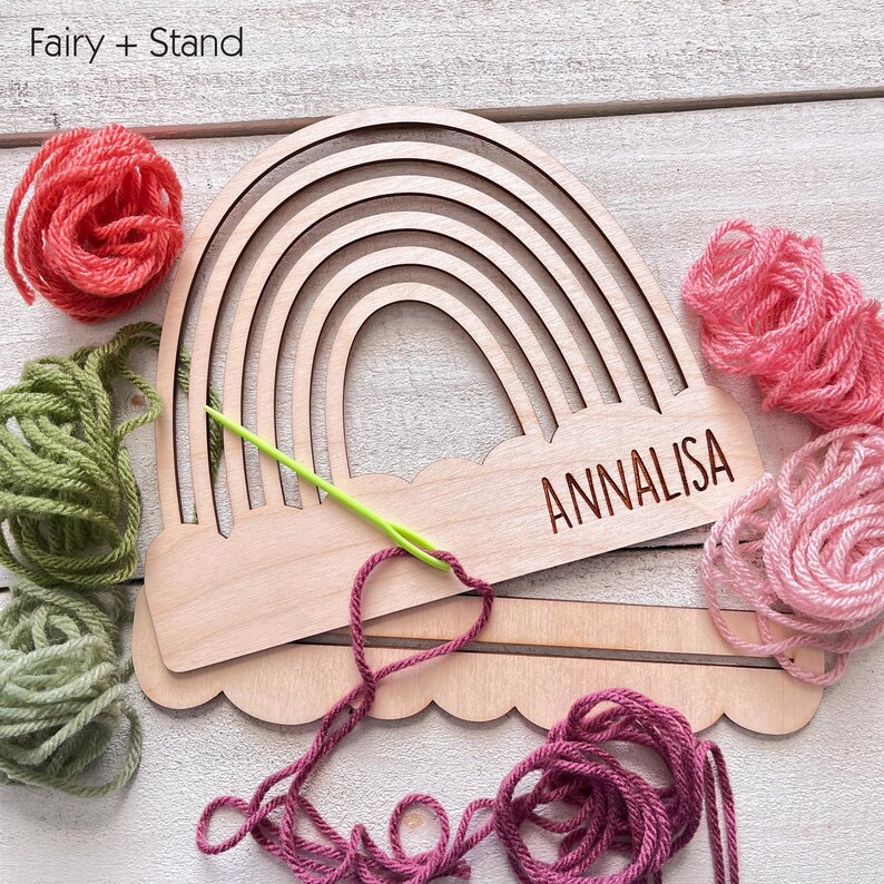 Pu&ograve; includere: Artigianato in legno a forma di arcobaleno con il nome "ANNALISA" inciso sulla base. Gomitoli di lana rosa, verde e rossa circondano l'oggetto, con un ago verde infilato con filo rosa. Il testo "Fairy + Stand" &egrave; nell'angolo in alto a sinistra.