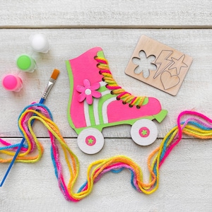 May include: A wooden roller skate cutout painted neon pink and lime green with a pink flower accent, featuring yellow and pink yarn laces, a paintbrush, three small paint pots, and a wooden stencil with flower, lightning, heart, and star shapes.