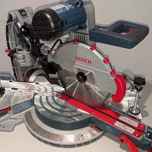 May include: A silver and blue Bosch miter saw with a red base and blade guard. The saw has a large circular blade and a handle for operation. The Bosch logo is visible on the blade guard.