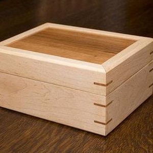 May include: A rectangular wooden box with a light-colored body and a darker wood top. The box features dovetail joints and is likely used for storage or display. The box is sitting on a dark wooden surface.