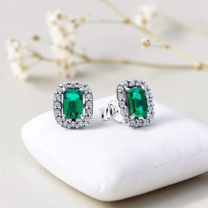 May include: Silver stud earrings featuring emerald-cut green gemstones, encircled by a halo of small, clear stones. The earrings are presented on a white surface.