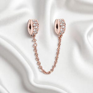 Puede incluir: Charm de oro rosa con dos extremos cilíndricos incrustados con cristales transparentes, unidos por una delicada cadena. El charm está diseñado para ser fijado a una pulsera o collar, añadiendo un toque de elegancia y brillo. Fondo blanco de tela sedosa.
