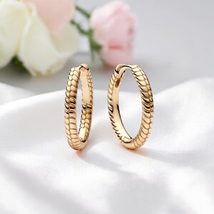 May include: A pair of gold-colored hoop earrings. The earrings have a textured, scale-like pattern. The background features white fabric and blurred flowers in pink and white.