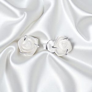 May include: A pair of white rose-shaped earrings with silver detailing. The earrings are displayed on a white satin surface, accentuating their delicate design. The floral theme adds a touch of elegance.